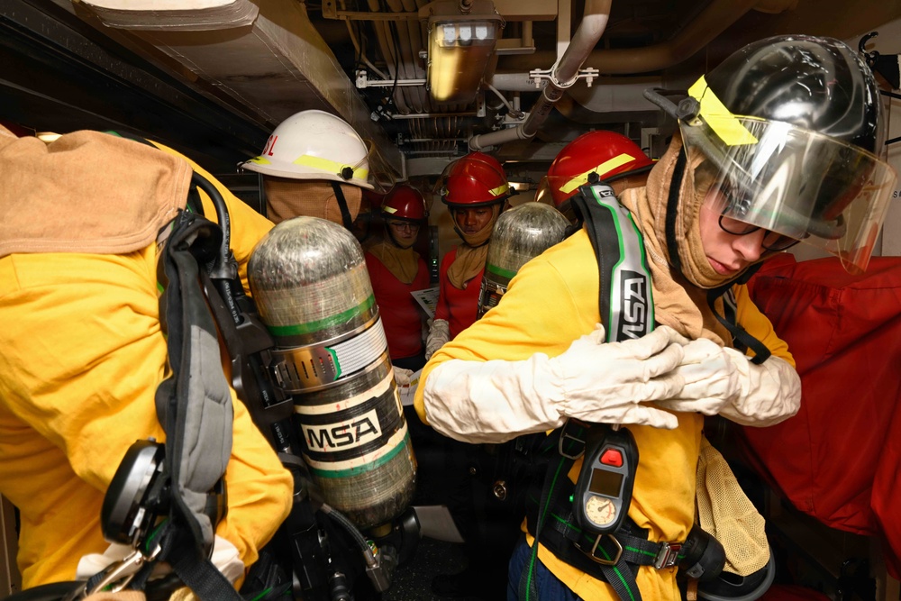 A group of personnel in firefighting or damage control gear are assembled in a confined shipboard compartment. They are wearing yellow fire-resistant suits, helmets of various colors, and MSA self-contained breathing apparatus (SCBA) tanks. The scene appears to be a training exercise or emergency response drill aboard a naval vessel.