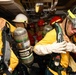 A group of personnel in firefighting or damage control gear are assembled in a confined shipboard compartment. They are wearing yellow fire-resistant suits, helmets of various colors, and MSA self-contained breathing apparatus (SCBA) tanks. The scene appears to be a training exercise or emergency response drill aboard a naval vessel.