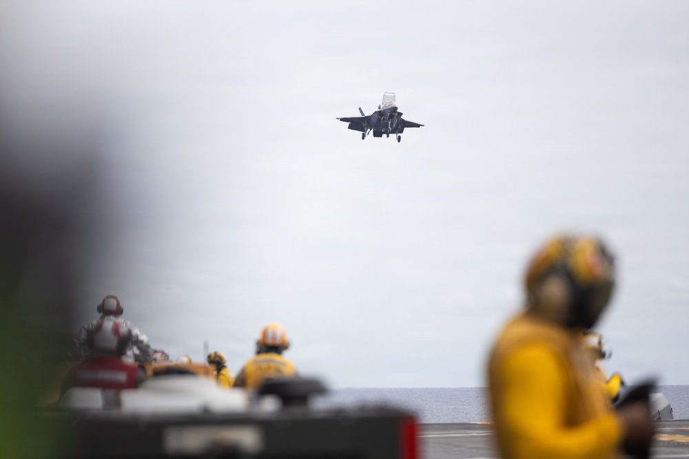 An F-35B fighter jet is captured in a short takeoff or vertical landing approach above the flight deck of what appears to be an amphibious assault ship or aircraft carrier. Multiple deck crew members wearing yellow flight deck jerseys and helmets are visible observing or managing operations. The image uses selective focus, with foreground and background elements intentionally blurred.