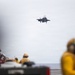 An F-35B fighter jet is captured in a short takeoff or vertical landing approach above the flight deck of what appears to be an amphibious assault ship or aircraft carrier. Multiple deck crew members wearing yellow flight deck jerseys and helmets are visible observing or managing operations. The image uses selective focus, with foreground and background elements intentionally blurred.