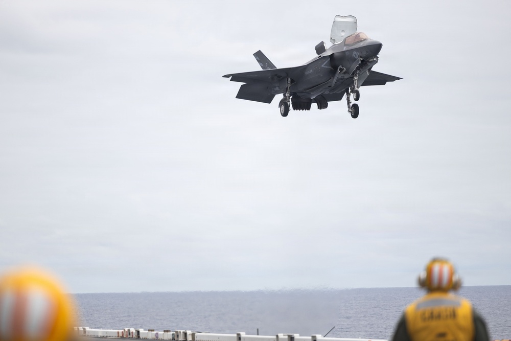 An F-35B Lightning II stealth fighter jet is performing a vertical or short takeoff/landing (VSTOL) approach over the deck of a naval vessel, likely an amphibious assault ship. Two deck crew members in yellow jerseys and helmets are visible on the flight deck observing the aircraft. The overcast sky and open ocean are visible in the background.