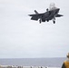 An F-35B Lightning II stealth fighter jet is performing a vertical or short takeoff/landing (VSTOL) approach over the deck of a naval vessel, likely an amphibious assault ship. Two deck crew members in yellow jerseys and helmets are visible on the flight deck observing the aircraft. The overcast sky and open ocean are visible in the background.