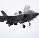 A U.S. Marines F-35B Lightning II stealth fighter jet is captured in flight with landing gear deployed and the lift fan door open on top of the fuselage, indicating a short takeoff or vertical landing (STOVL) configuration. The aircraft bears U.S. Marine Corps markings and is photographed against an overcast sky. The tail code and fuselage markings suggest it belongs to a Marine Fighter Attack Training Squadron.