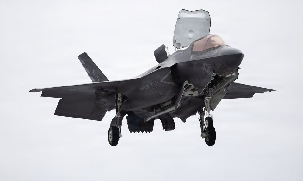 A U.S. military F-35B Lightning II stealth fighter jet is captured in flight with landing gear fully deployed, suggesting an approach to landing or a short takeoff/vertical landing (STOVL) maneuver. The aircraft is photographed head-on against an overcast, grey sky. The lift fan door on top of the fuselage is open, which is characteristic of the F-35B variant used by the U.S. Marine Corps.