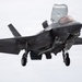 A U.S. military F-35B Lightning II stealth fighter jet is captured in flight with landing gear fully deployed, suggesting an approach to landing or a short takeoff/vertical landing (STOVL) maneuver. The aircraft is photographed head-on against an overcast, grey sky. The lift fan door on top of the fuselage is open, which is characteristic of the F-35B variant used by the U.S. Marine Corps.