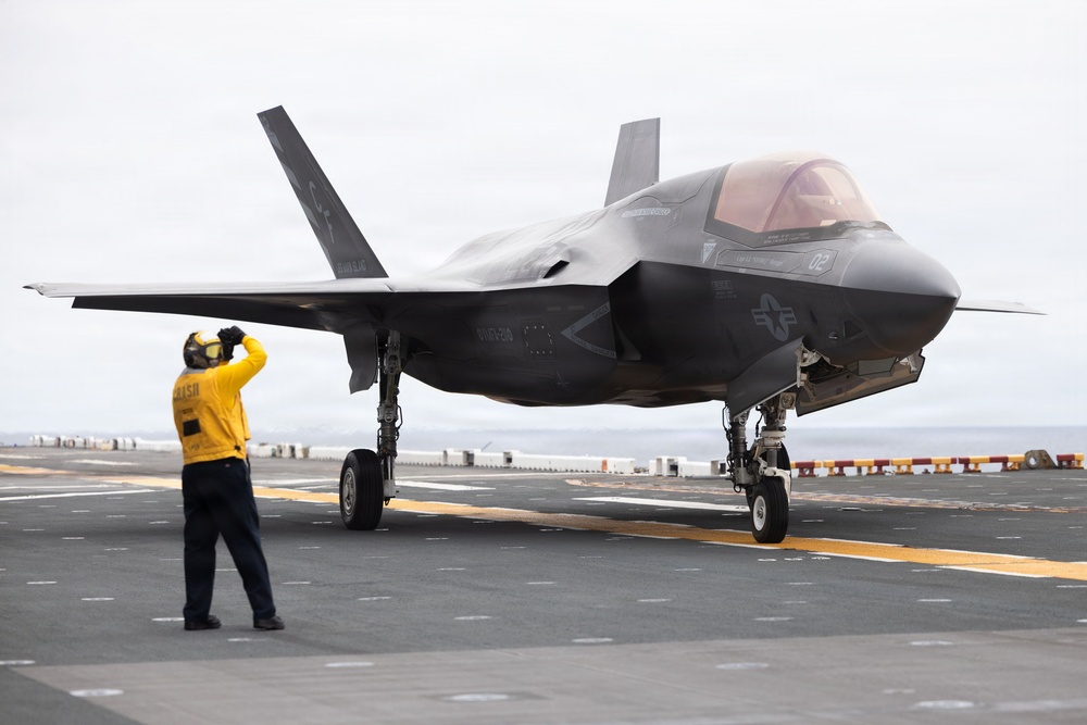 A dark-colored stealth fighter jet, identifiable as an F-35B, is positioned on the flight deck of an aircraft carrier. A flight deck crew member in a yellow jersey is signaling toward the aircraft. The scene appears to be during taxi or pre-launch operations aboard a naval vessel.