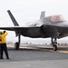 A dark-colored stealth fighter jet, identifiable as an F-35B, is positioned on the flight deck of an aircraft carrier. A flight deck crew member in a yellow jersey is signaling toward the aircraft. The scene appears to be during taxi or pre-launch operations aboard a naval vessel.