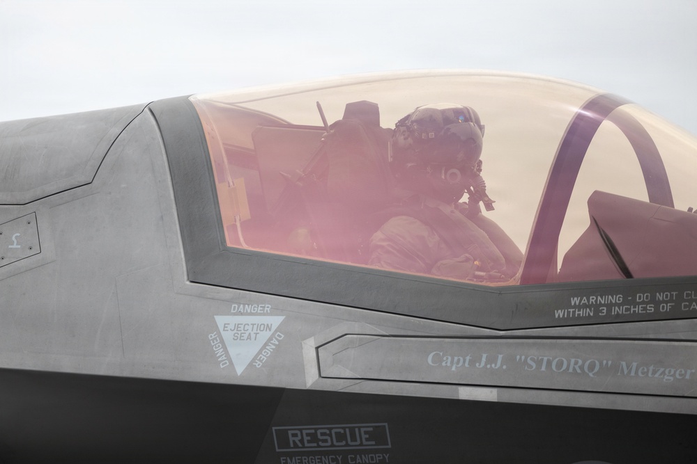 A close-up photograph of a military fighter jet cockpit, identified as belonging to Capt. J.J. 'STORQ' Metzger. A pilot wearing a flight helmet and oxygen mask is seated inside the cockpit beneath a tinted canopy. The aircraft displays standard safety markings including ejection seat warnings and rescue instructions.