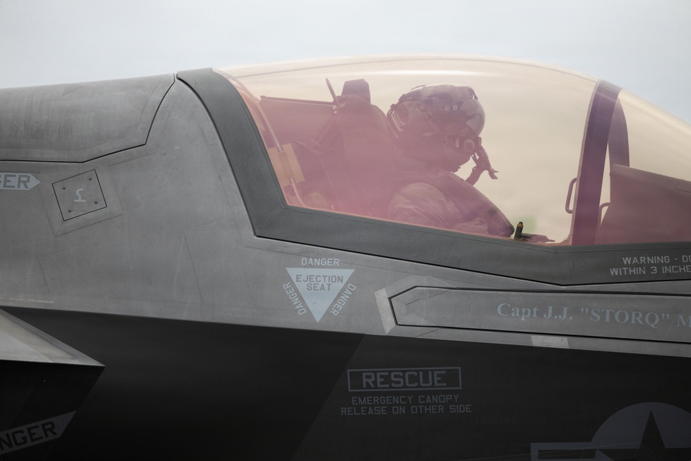 A military pilot is seated in the cockpit of what appears to be an F-35 Lightning II stealth fighter jet. The pilot is wearing a flight helmet and oxygen mask, visible through the tinted canopy. Several safety and rescue markings are clearly visible on the aircraft's fuselage.