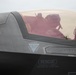A military pilot is seated in the cockpit of what appears to be an F-35 Lightning II stealth fighter jet. The pilot is wearing a flight helmet and oxygen mask, visible through the tinted canopy. Several safety and rescue markings are clearly visible on the aircraft's fuselage.