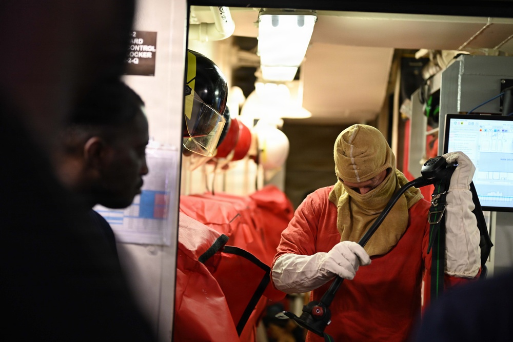 The image shows personnel in a confined interior space, likely aboard a naval vessel, donning firefighting or damage control protective gear. At least two individuals are visible, with one fully suited in red protective clothing, a tan flash hood, white gloves, and holding breathing apparatus equipment. A sign partially reading 'DAMAGE CONTROL LOCKER' is visible on the wall, confirming a naval damage control context.