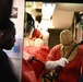The image shows personnel in a confined interior space, likely aboard a naval vessel, donning firefighting or damage control protective gear. At least two individuals are visible, with one fully suited in red protective clothing, a tan flash hood, white gloves, and holding breathing apparatus equipment. A sign partially reading 'DAMAGE CONTROL LOCKER' is visible on the wall, confirming a naval damage control context.