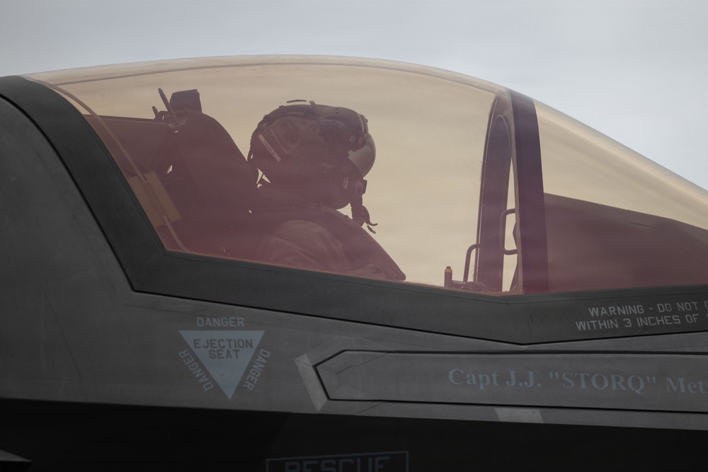 A military pilot is seated in the cockpit of what appears to be an F-35 fighter jet, visible through the tinted canopy. The aircraft fuselage displays safety markings and a pilot name placard reading 'Capt J.J. "STORQ" Me...' (partially visible). The image is taken from an exterior angle close to the cockpit.