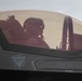A military pilot is seated in the cockpit of what appears to be an F-35 fighter jet, visible through the tinted canopy. The aircraft fuselage displays safety markings and a pilot name placard reading 'Capt J.J. "STORQ" Me...' (partially visible). The image is taken from an exterior angle close to the cockpit.