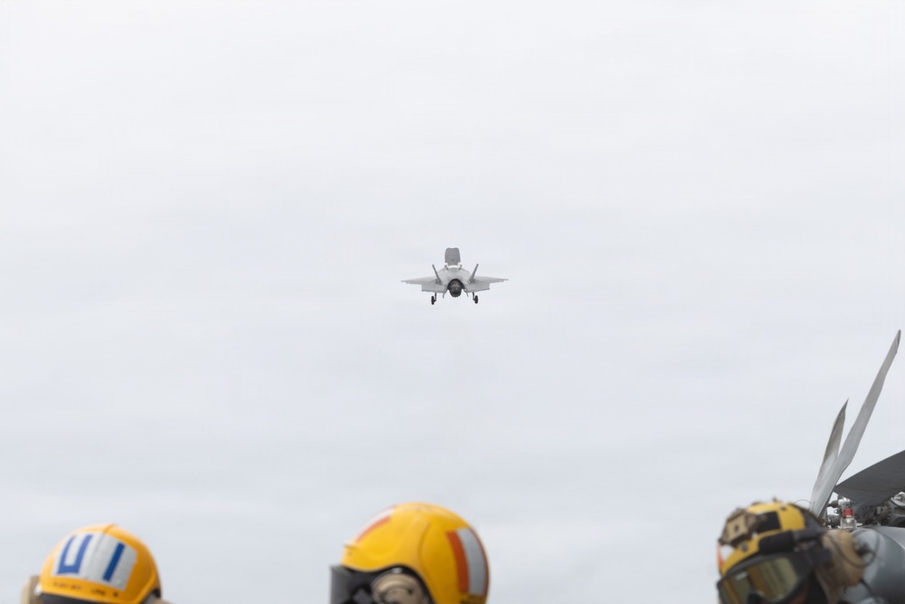 A military jet aircraft, consistent with an F-35B, is captured in flight approaching what appears to be a naval vessel, likely an aircraft carrier or amphibious assault ship. Several deck crew members wearing yellow helmets and protective gear are visible in the foreground observing the aircraft. The overcast sky and deck environment suggest active flight operations at sea.