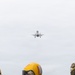 A military jet aircraft, consistent with an F-35B, is captured in flight approaching what appears to be a naval vessel, likely an aircraft carrier or amphibious assault ship. Several deck crew members wearing yellow helmets and protective gear are visible in the foreground observing the aircraft. The overcast sky and deck environment suggest active flight operations at sea.