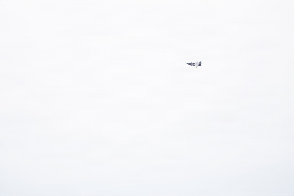 A single military jet aircraft is visible flying at high altitude against a uniformly overcast, pale grey-white sky. The aircraft appears small and distant, positioned in the upper-right quadrant of the frame. No ground features or other objects are present in the image.