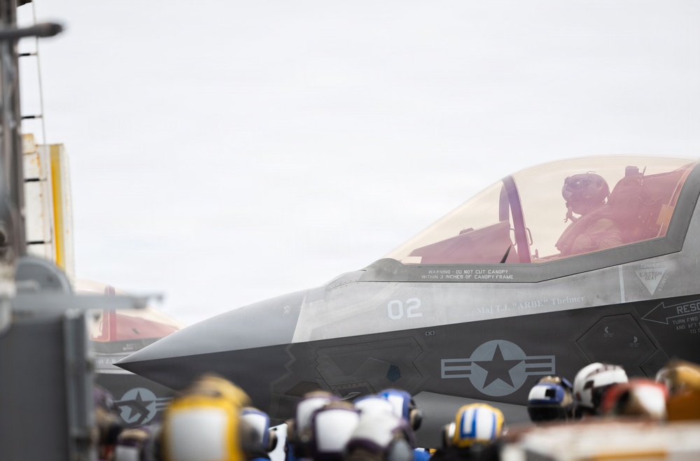 A military jet aircraft, identified as aircraft number '02', is positioned on what appears to be an aircraft carrier flight deck. A pilot is seated in the cockpit wearing a flight helmet. Ground crew personnel wearing various colored helmets are visible in the foreground.
