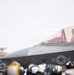 A military jet aircraft, identified as aircraft number '02', is positioned on what appears to be an aircraft carrier flight deck. A pilot is seated in the cockpit wearing a flight helmet. Ground crew personnel wearing various colored helmets are visible in the foreground.