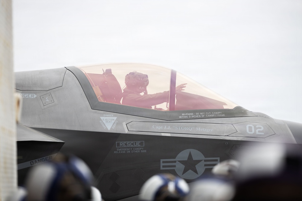 A military pilot is seated in the cockpit of an F-35 fighter jet, designated aircraft number 02, assigned to Capt. J.J. 'STORQ' Metzger. The aircraft displays standard U.S. military markings including a roundel and safety/rescue stenciling. Ground crew personnel wearing helmets are visible in the foreground, suggesting pre-flight or taxiing operations.