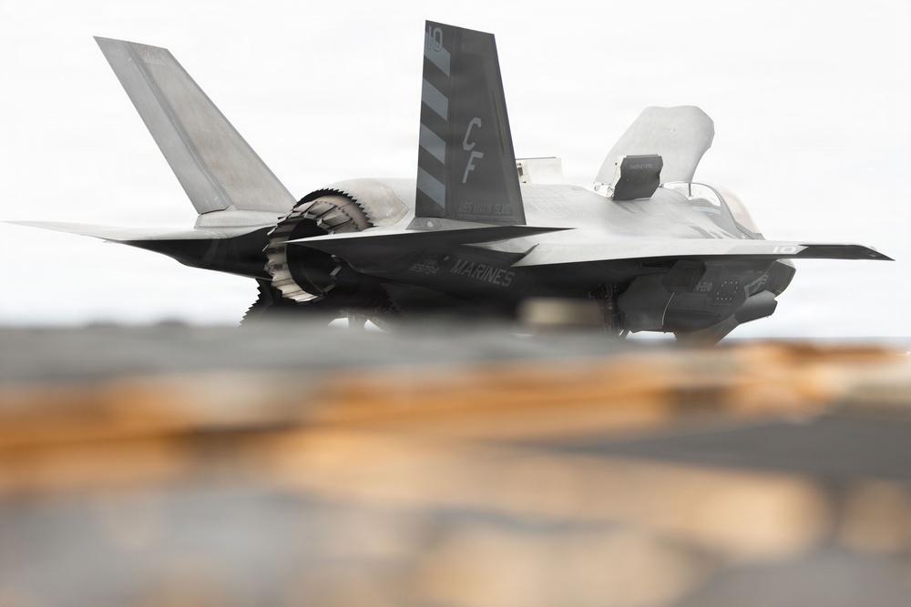 A U.S. Marines F-35B stealth fighter jet is captured in a low-angle shot, appearing to be on or just above a ship's deck, likely an amphibious assault ship. The aircraft displays 'MARINES' markings on the fuselage and tail code 'CF' with chevron stripes. The lift fan opening is visible on top of the fuselage, consistent with the F-35B short takeoff/vertical landing (STOVL) variant.