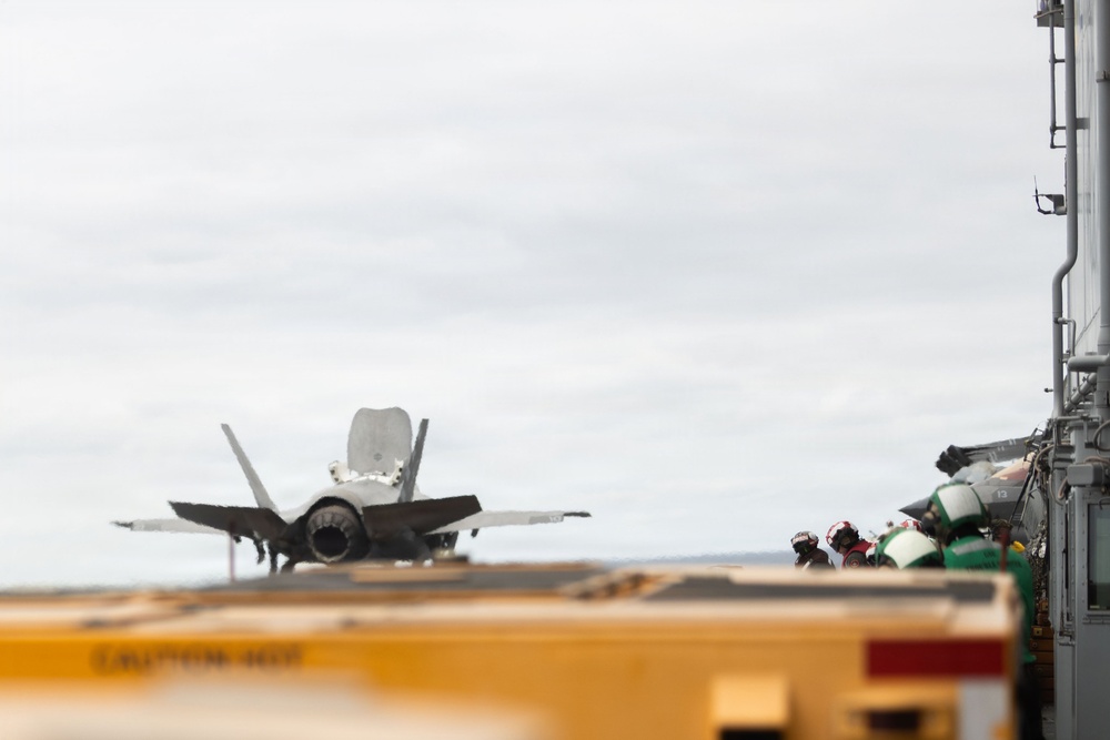 An F-35 fighter jet is positioned on the flight deck of what appears to be a naval aircraft carrier, viewed from the rear/aft angle. Several deck crew personnel wearing helmets and protective gear are visible along the island structure on the right side of the frame. The image captures a carrier flight deck operation under overcast skies.