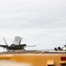 An F-35 fighter jet is positioned on the flight deck of what appears to be a naval aircraft carrier, viewed from the rear/aft angle. Several deck crew personnel wearing helmets and protective gear are visible along the island structure on the right side of the frame. The image captures a carrier flight deck operation under overcast skies.