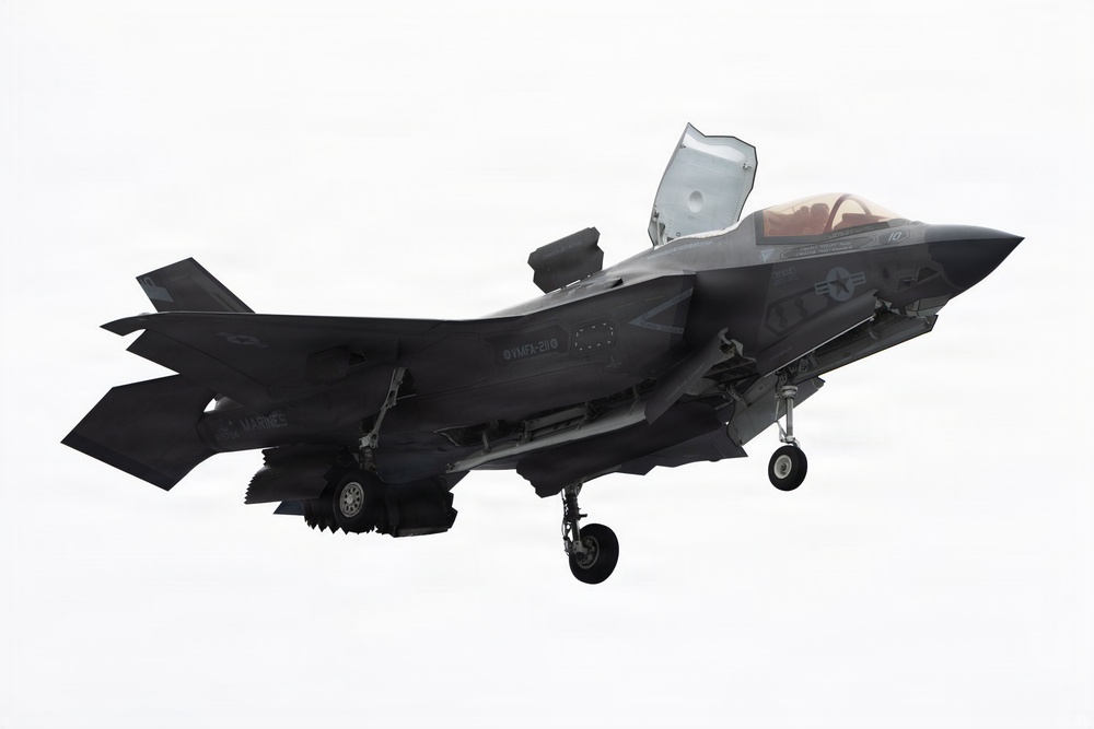 A U.S. Marines F-35B Lightning II stealth fighter jet is captured in low-speed flight with landing gear deployed and the lift fan door open on top of the fuselage, indicating a short takeoff or vertical landing (STOVL) configuration. The aircraft bears U.S. Marine Corps markings and unit designation 'VMFA-211' on the fuselage. The image is taken against a pale, overcast sky background.