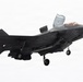 A U.S. Marines F-35B Lightning II stealth fighter jet is captured in low-speed flight with landing gear deployed and the lift fan door open on top of the fuselage, indicating a short takeoff or vertical landing (STOVL) configuration. The aircraft bears U.S. Marine Corps markings and unit designation 'VMFA-211' on the fuselage. The image is taken against a pale, overcast sky background.