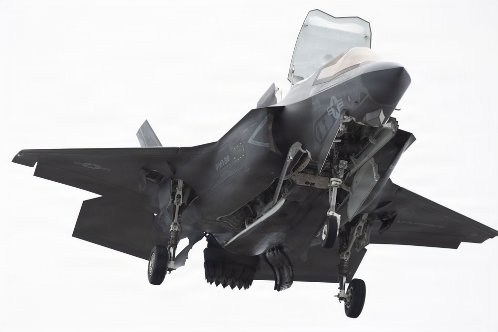 A dark gray stealth fighter jet, identifiable as an F-35B Lightning II, is captured in flight with landing gear fully deployed, suggesting it is in a landing or low-speed hover configuration. The aircraft is photographed from a slightly below-front angle against a bright, overcast white sky. The lift fan door and other panels appear open, consistent with the F-35B's Short Take-Off and Vertical Landing (STOVL) capability.