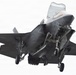 A dark gray stealth fighter jet, identifiable as an F-35B Lightning II, is captured in flight with landing gear fully deployed, suggesting it is in a landing or low-speed hover configuration. The aircraft is photographed from a slightly below-front angle against a bright, overcast white sky. The lift fan door and other panels appear open, consistent with the F-35B's Short Take-Off and Vertical Landing (STOVL) capability.