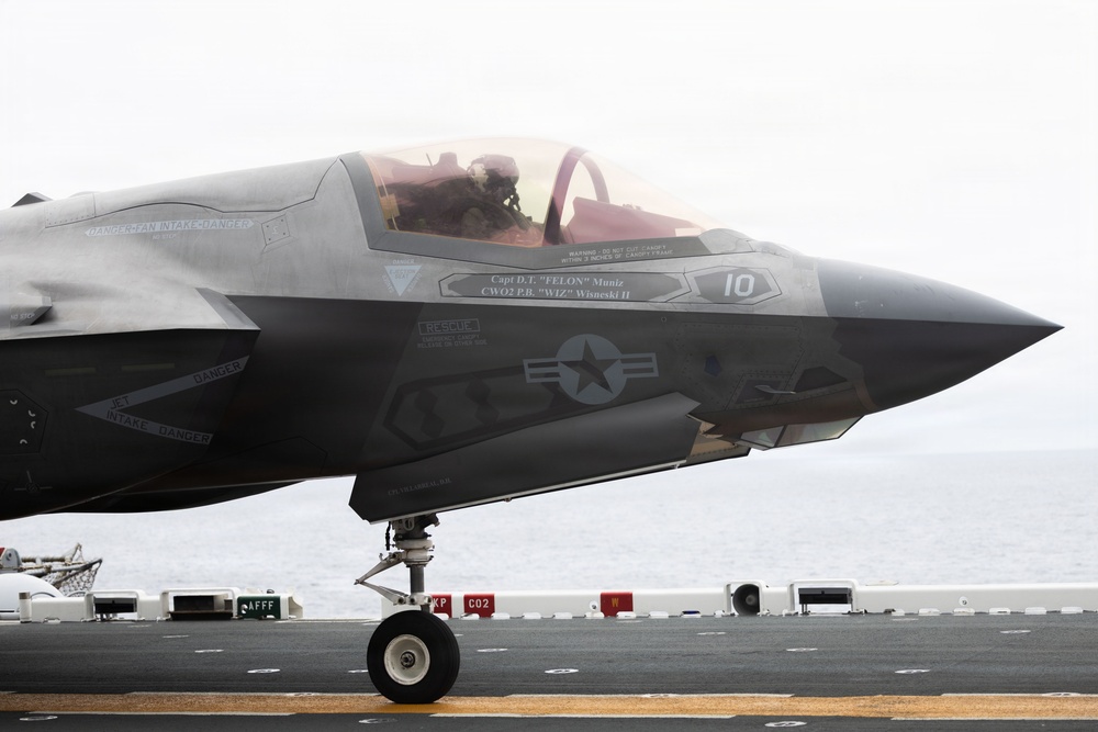 A U.S. Marine Corps F-35B Lightning II stealth fighter jet is positioned on the flight deck of an aircraft carrier at sea. The aircraft bears the number '10' and crew names 'Capt D.T. FELON Muniz' and 'CWO2 P.B. WIZ Wisneski II' stenciled below the cockpit. A pilot is visible in the cockpit, and the ocean is visible in the background.