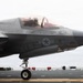 A U.S. Marine Corps F-35B Lightning II stealth fighter jet is positioned on the flight deck of an aircraft carrier at sea. The aircraft bears the number '10' and crew names 'Capt D.T. FELON Muniz' and 'CWO2 P.B. WIZ Wisneski II' stenciled below the cockpit. A pilot is visible in the cockpit, and the ocean is visible in the background.