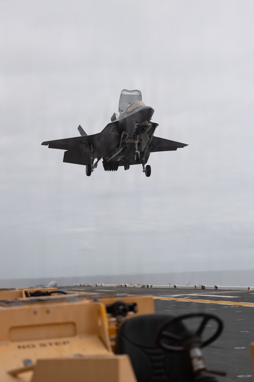 An F-35B Lightning II stealth fighter jet is captured in a vertical or short takeoff/landing (VSTOL) maneuver above the flight deck of an amphibious assault ship. The aircraft's landing gear is deployed and the lift fan door is open, indicating a vertical landing or hover approach. Ground support equipment and deck markings are visible in the foreground.
