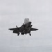 An F-35B Lightning II stealth fighter jet is captured in a vertical or short takeoff/landing (VSTOL) maneuver above the flight deck of an amphibious assault ship. The aircraft's landing gear is deployed and the lift fan door is open, indicating a vertical landing or hover approach. Ground support equipment and deck markings are visible in the foreground.
