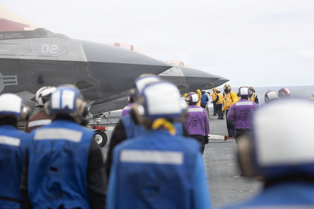 A stealth fighter jet, identified as aircraft number '02' and consistent with an F-35 variant, is positioned on the flight deck of an aircraft carrier. Deck crew members in color-coded jerseys and helmets are visible performing flight deck operations. The scene depicts a standard carrier air wing operation with multiple personnel fulfilling specialized roles.