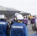 A stealth fighter jet, identified as aircraft number '02' and consistent with an F-35 variant, is positioned on the flight deck of an aircraft carrier. Deck crew members in color-coded jerseys and helmets are visible performing flight deck operations. The scene depicts a standard carrier air wing operation with multiple personnel fulfilling specialized roles.