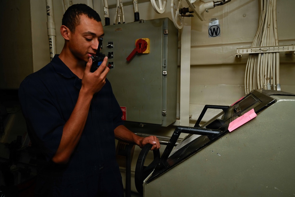 A young man in a dark navy blue uniform is using a handheld radio/walkie-talkie while standing at what appears to be a control console or equipment station aboard a military vessel. He is holding a handle on the console with his other hand while communicating. The setting is an interior compartment of what appears to be a naval ship, with electrical panels, cable runs, and military-grade equipment visible.