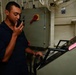 A young man in a dark navy blue uniform is using a handheld radio/walkie-talkie while standing at what appears to be a control console or equipment station aboard a military vessel. He is holding a handle on the console with his other hand while communicating. The setting is an interior compartment of what appears to be a naval ship, with electrical panels, cable runs, and military-grade equipment visible.