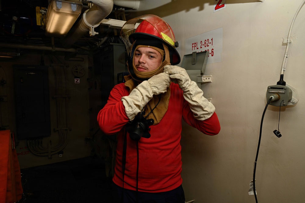 A young person is donning firefighting gear inside what appears to be a naval vessel. They are wearing a red firefighting helmet with a face shield, a tan fire-resistant hood, white firefighting gloves, and a red long-sleeve shirt. The setting shows industrial naval interior features including pipes, conduits, and a labeled panel marked 'FPL 2-62-1'.