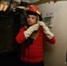 A young person is donning firefighting gear inside what appears to be a naval vessel. They are wearing a red firefighting helmet with a face shield, a tan fire-resistant hood, white firefighting gloves, and a red long-sleeve shirt. The setting shows industrial naval interior features including pipes, conduits, and a labeled panel marked 'FPL 2-62-1'.