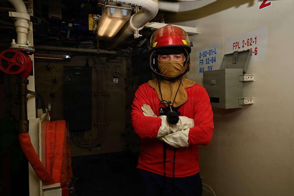 A person wearing firefighting/damage control gear stands with arms crossed inside what appears to be a naval vessel. They are equipped with a red helmet, flash hood, white gloves, and a red long-sleeve shirt, consistent with U.S. Navy damage control attire. The surrounding environment shows shipboard infrastructure including pipes, valves, electrical panels, and compartment designation placards.