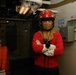 A person wearing firefighting/damage control gear stands with arms crossed inside what appears to be a naval vessel. They are equipped with a red helmet, flash hood, white gloves, and a red long-sleeve shirt, consistent with U.S. Navy damage control attire. The surrounding environment shows shipboard infrastructure including pipes, valves, electrical panels, and compartment designation placards.