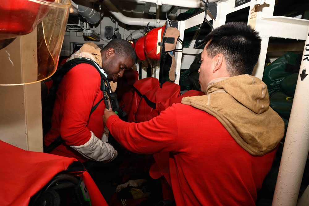 Two individuals are inside a confined shipboard compartment, with one person in a red shirt assisting the other in donning or adjusting what appears to be a survival or immersion suit. The setting is clearly a naval vessel interior, characterized by pipes, brackets, and stowed equipment. The interaction suggests a training exercise or emergency preparedness drill.