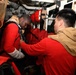 Two individuals are inside a confined shipboard compartment, with one person in a red shirt assisting the other in donning or adjusting what appears to be a survival or immersion suit. The setting is clearly a naval vessel interior, characterized by pipes, brackets, and stowed equipment. The interaction suggests a training exercise or emergency preparedness drill.