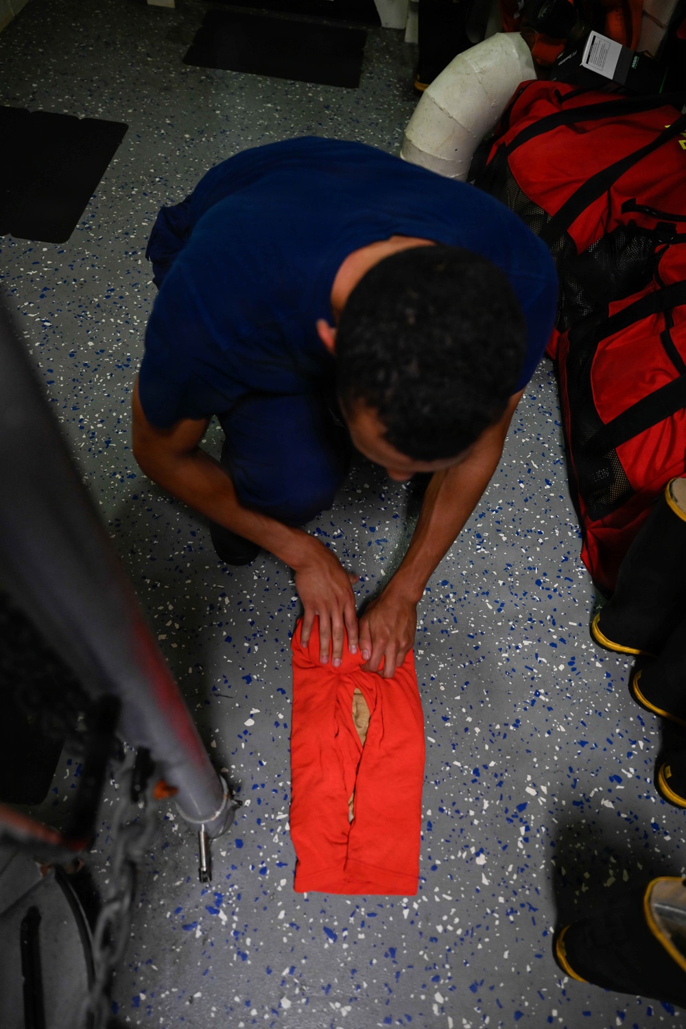 A person wearing a blue shirt is kneeling on a speckled gray floor, pressing their hands down on a red garment or fabric item that appears to have a tear or hole in it. The setting appears to be an indoor facility, possibly a training or emergency services environment, given the presence of red gear bags and rescue equipment nearby. The overhead angle provides a clear top-down view of the activity.