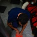 A person wearing a blue shirt is kneeling on a speckled gray floor, pressing their hands down on a red garment or fabric item that appears to have a tear or hole in it. The setting appears to be an indoor facility, possibly a training or emergency services environment, given the presence of red gear bags and rescue equipment nearby. The overhead angle provides a clear top-down view of the activity.