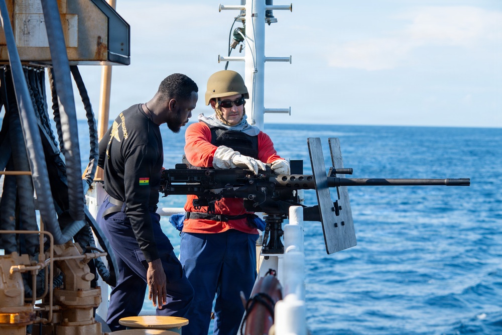 Two individuals are on the deck of a naval or coast guard vessel at sea, examining a mounted heavy machine gun. One person wearing a helmet, body armor, red long-sleeve shirt, and gloves appears to be demonstrating or instructing the other on the weapon system. The setting suggests a maritime training or familiarization exercise.