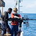 Two individuals are on the deck of a naval or coast guard vessel at sea, examining a mounted heavy machine gun. One person wearing a helmet, body armor, red long-sleeve shirt, and gloves appears to be demonstrating or instructing the other on the weapon system. The setting suggests a maritime training or familiarization exercise.