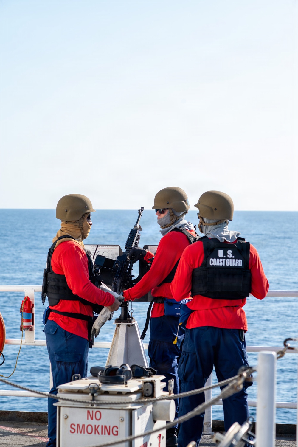 Three U.S. Coast Guard personnel are stationed on the deck of a vessel at sea, gathered around a mounted weapon system. They are wearing red long-sleeve shirts, navy blue pants, tactical vests, ballistic helmets, and face coverings. The scene appears to depict a weapons training or readiness drill conducted on open water.